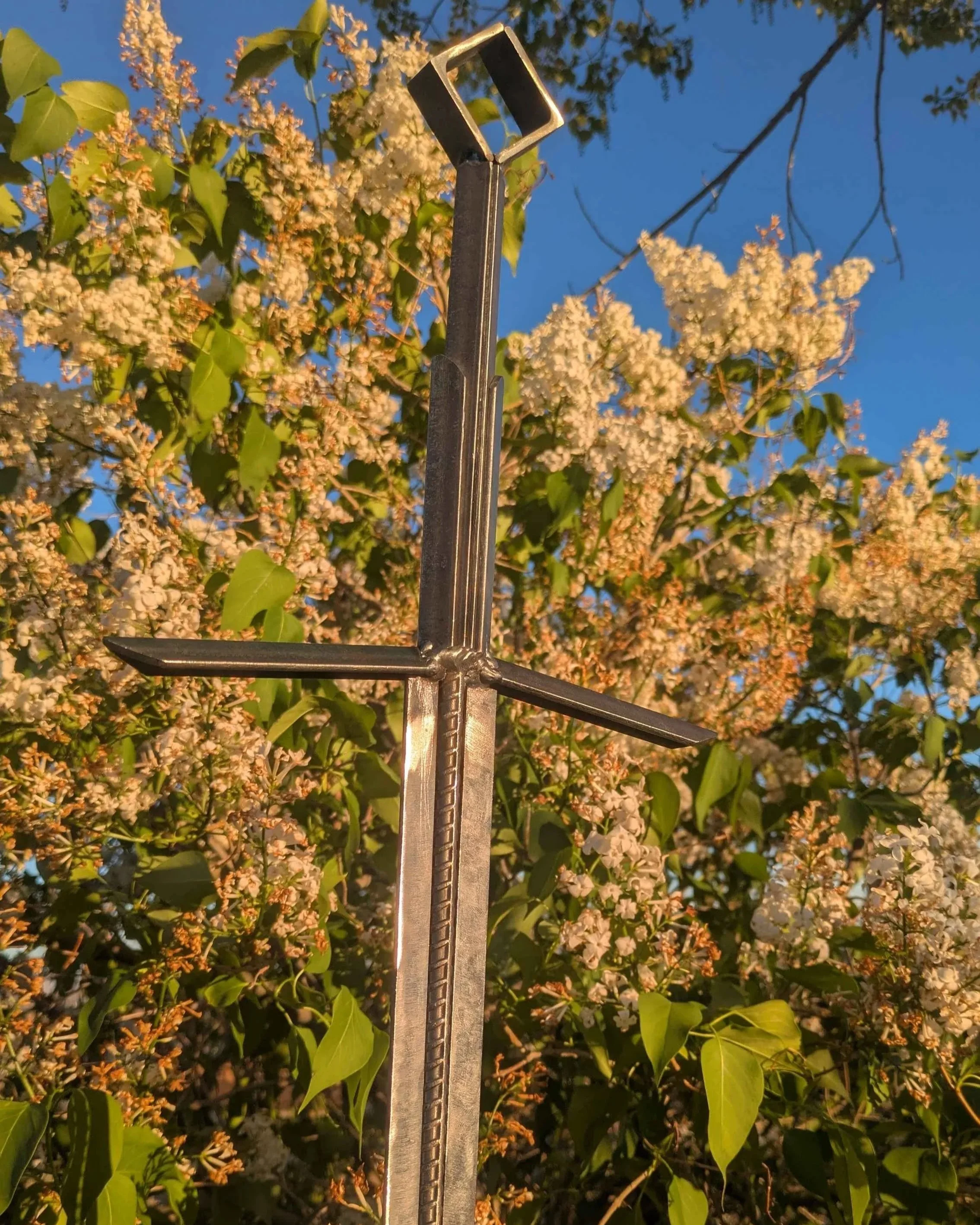 The hilt of a metal art sword made from industrial style materials, planted into the ground in front of a bush.