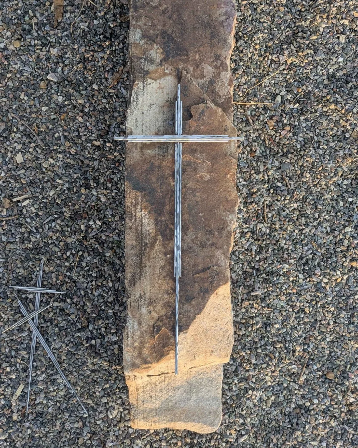 A large metal art cross made from long pieces of polished metal rod, with a long spike on the end for planting in a front yard.