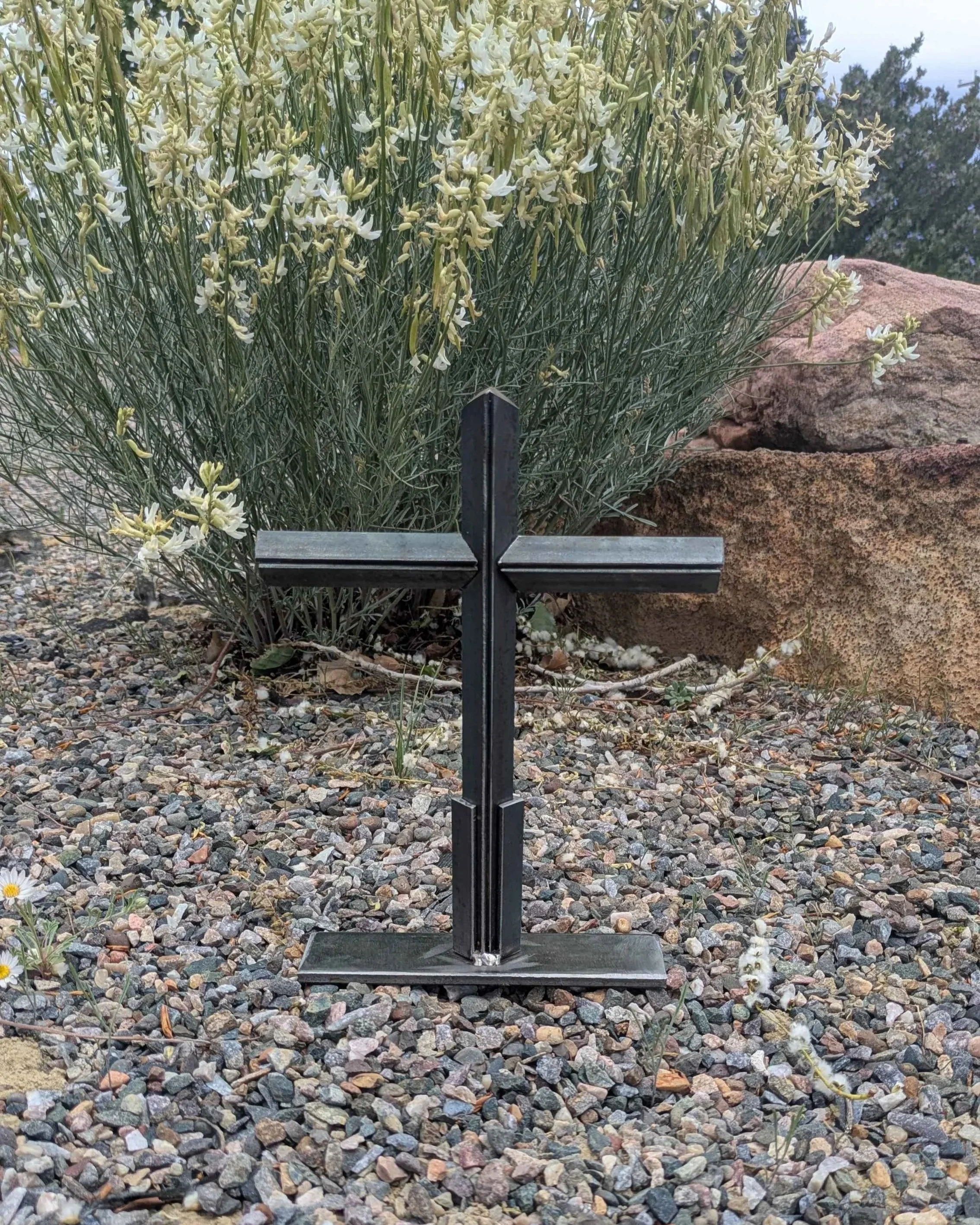 A metal art cross made from small pieces of angle iron placed on a stand, sitting upon gravel in front of a bush.