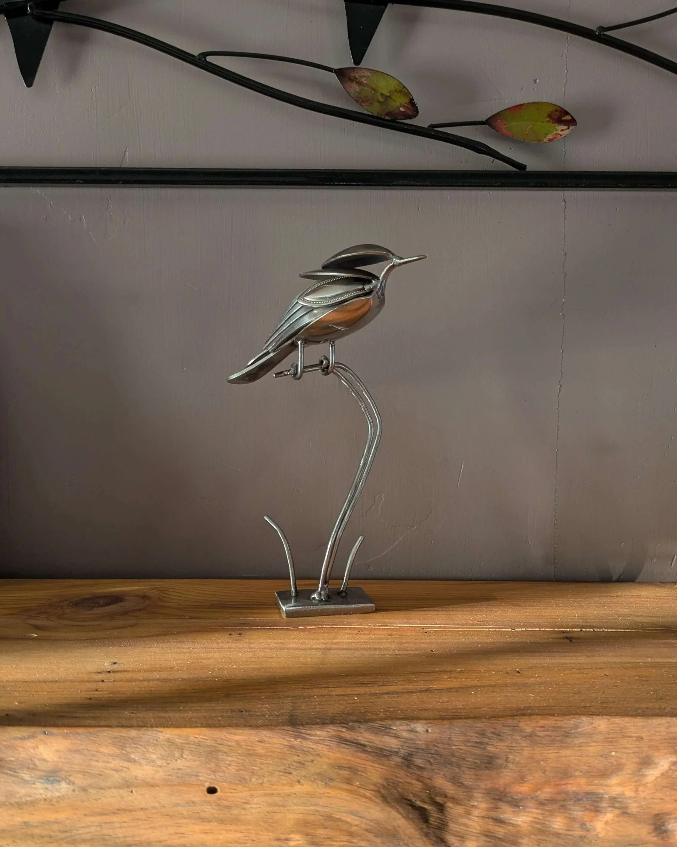 A handmade metal art bird sculpture made from recycled silverware sitting on a wooden shelf, resulting in a great piece of home decor.