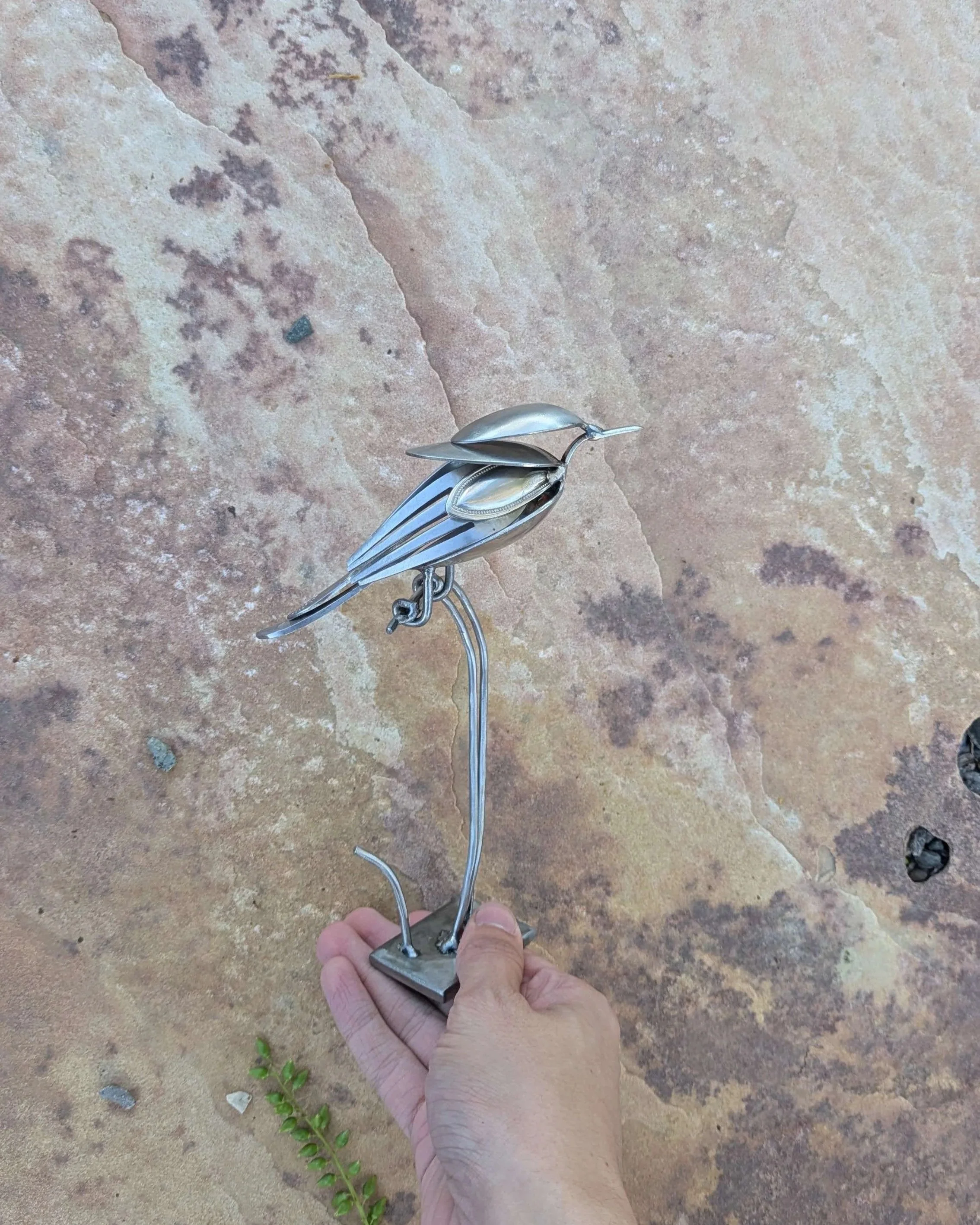 A handmade metal art bird sculpture made from recycled silverware sitting on a wooden shelf, resulting in a great piece of home decor.