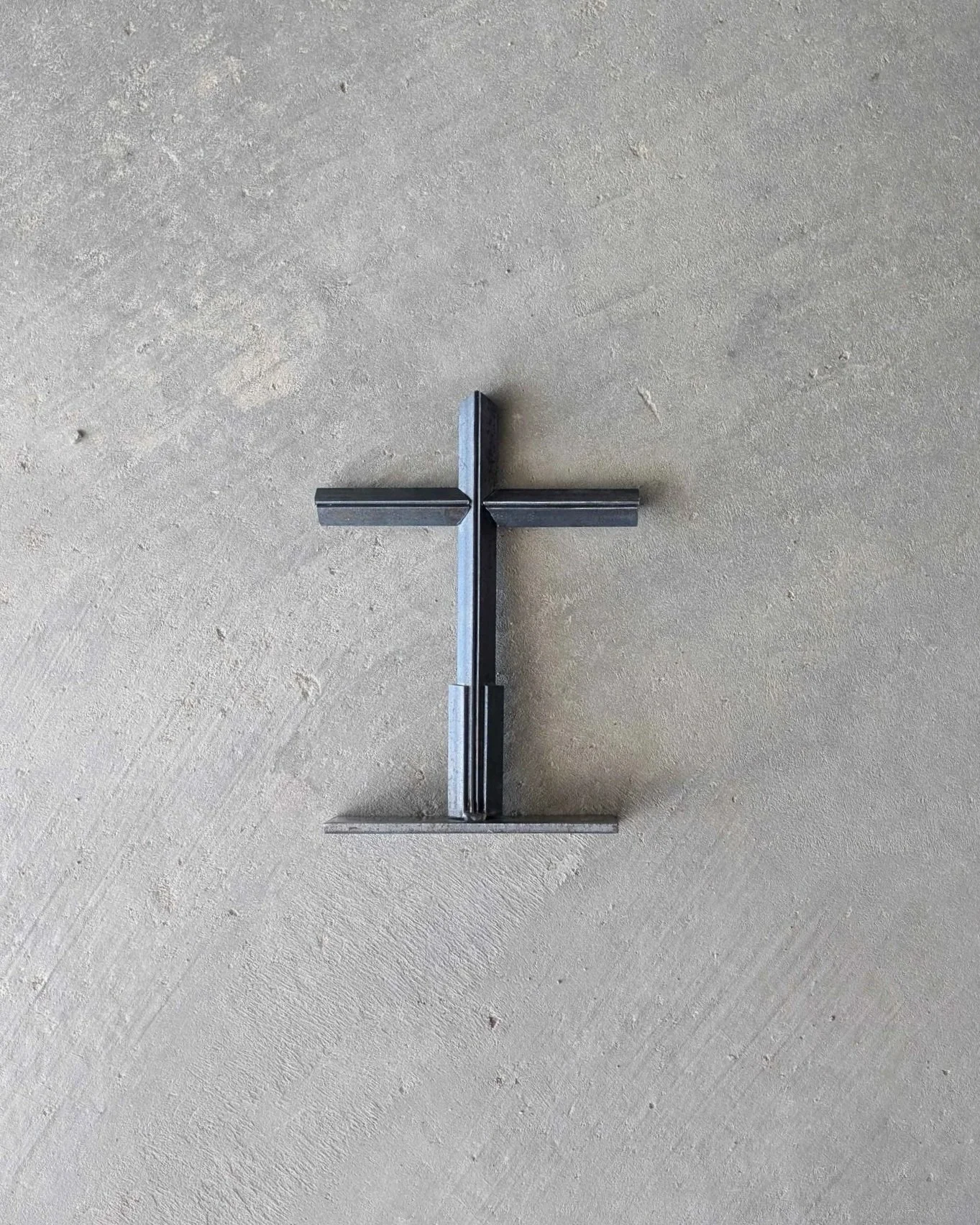 A metal art cross made from small pieces of angle iron placed on a stand, laying upon a slab of concrete..