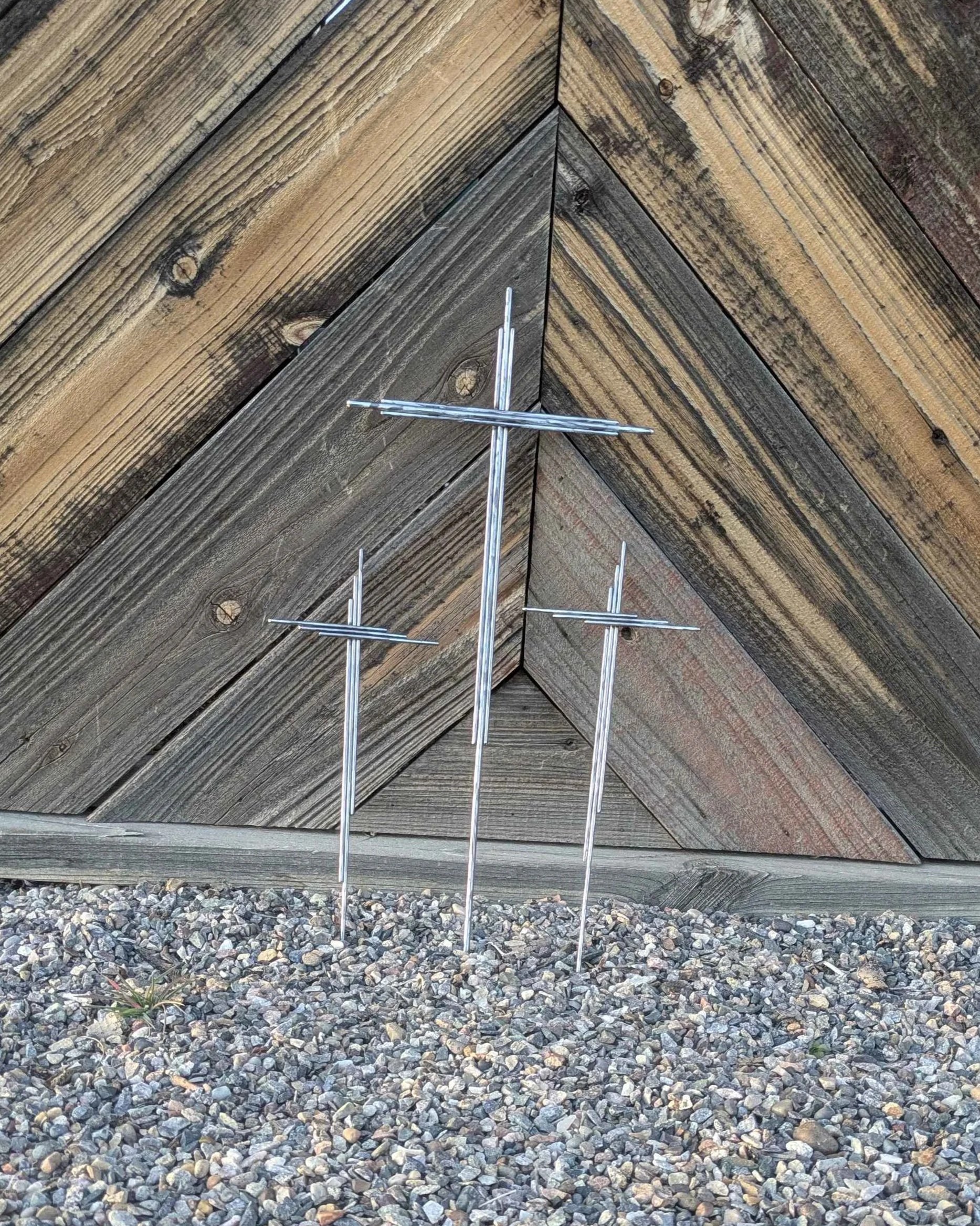 A large metal art cross made from long pieces of polished metal rod, with a long spike on the end for planting in a front yard.