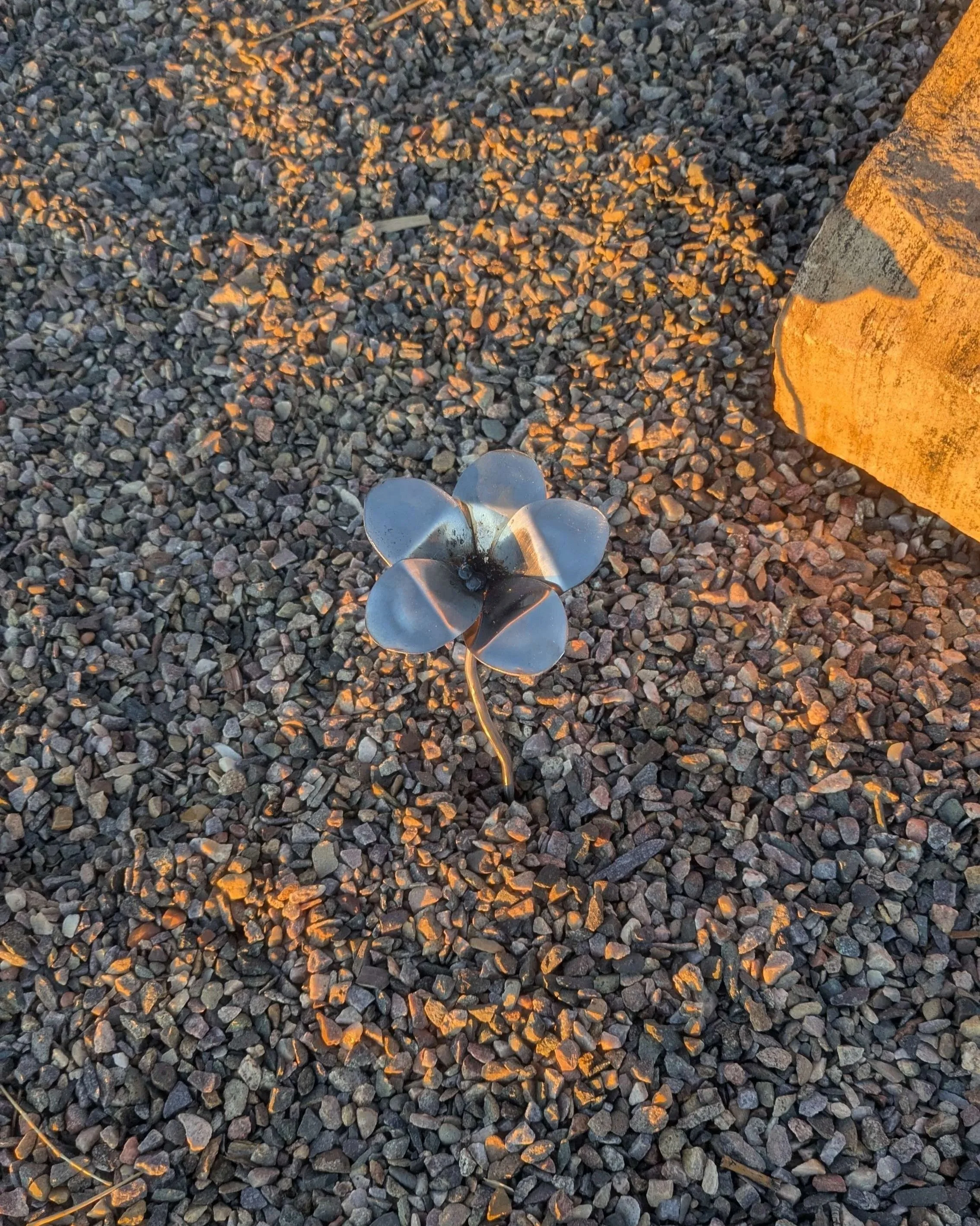 A metal art flower made from bent spoon heads and metal rod planted in gravel.