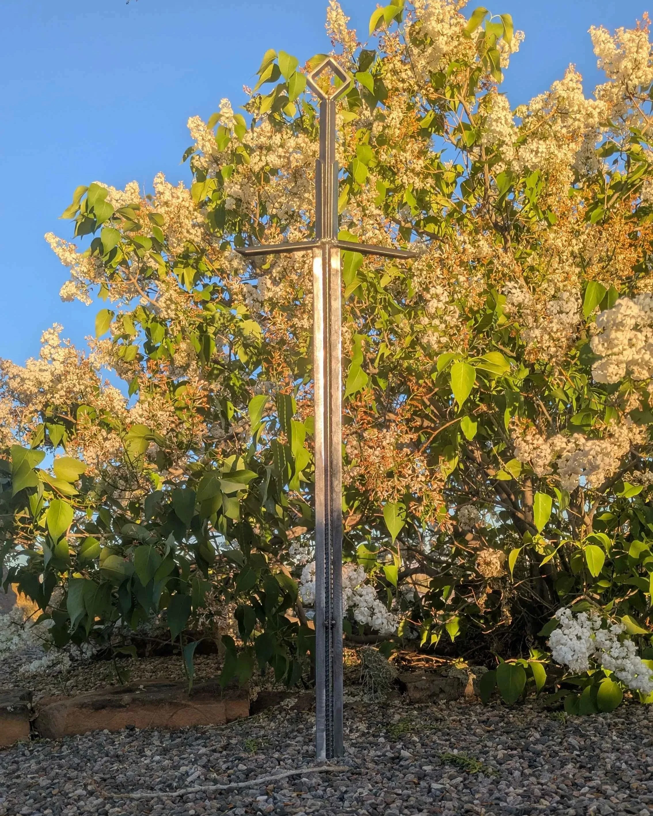 A metal art sword made from industrial style materials, planted into the ground in front of a bush.