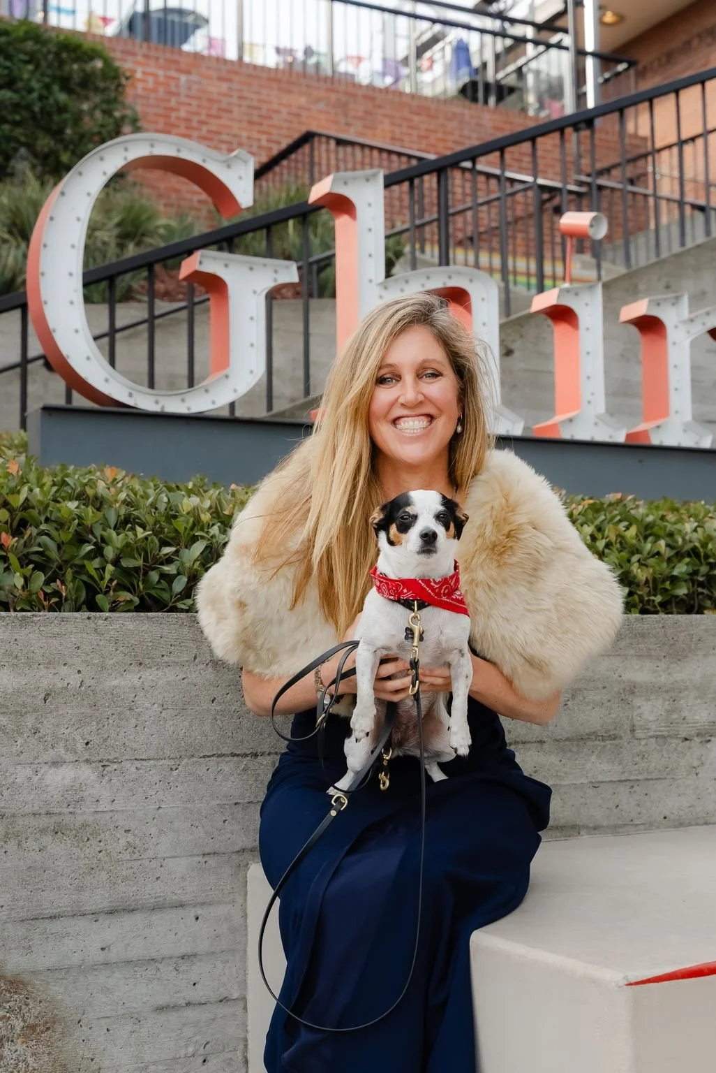 A real estate agent sitting on a step at Ghirardelli Square holding a small dog with a red bandana.