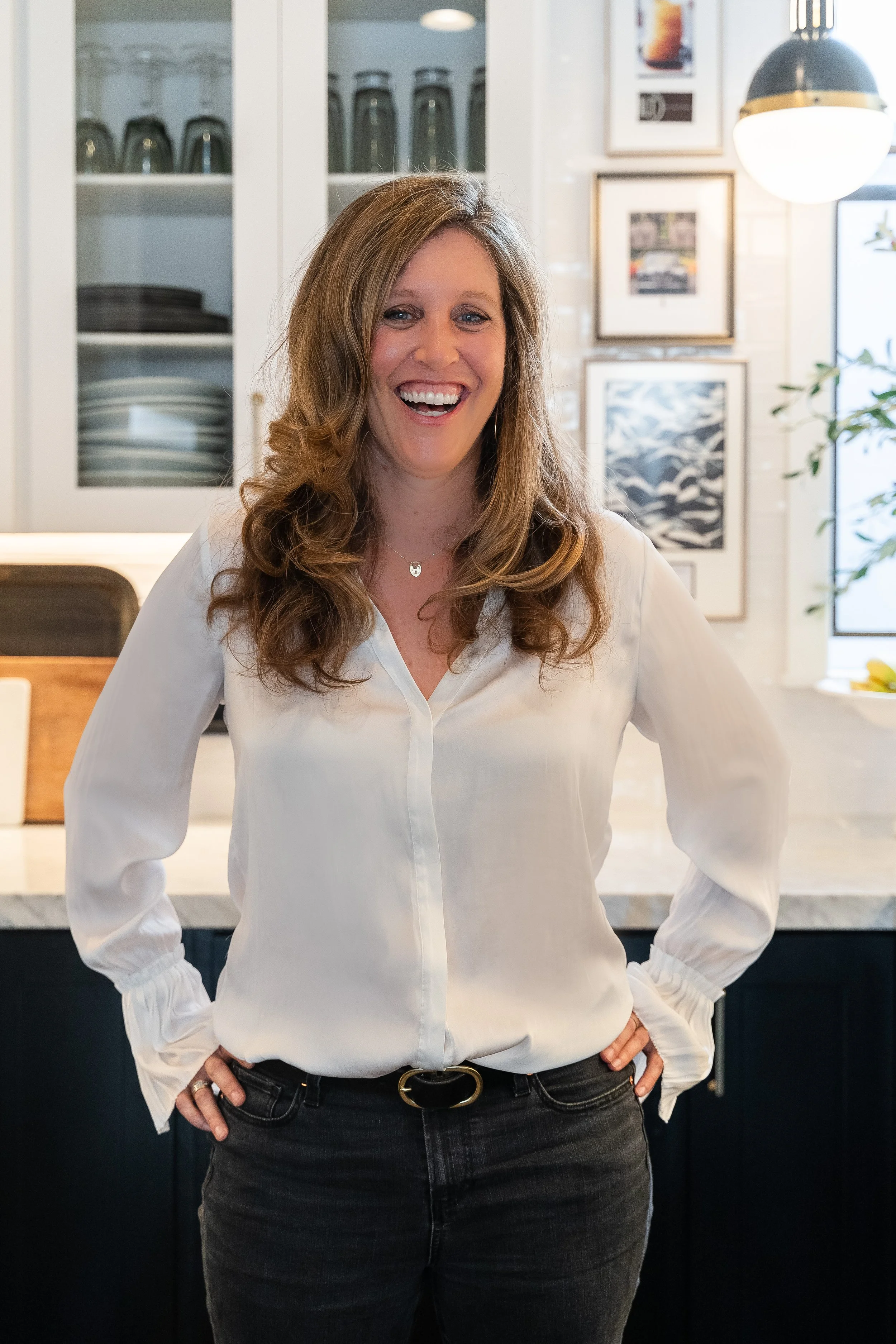 A woman with long wavy brown hair, smiling, wearing a white blouse and dark jeans, standing in a modern kitchen with framed artwork on the wall and a black and gold pendant light