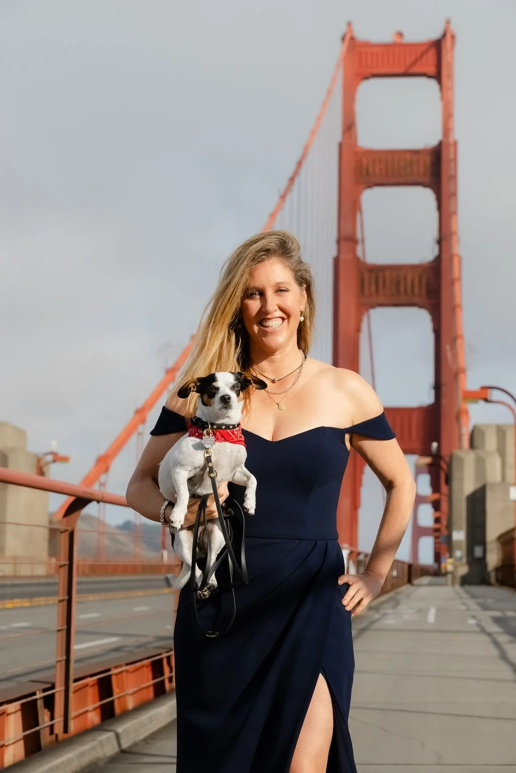 A real estate agent in a dark blue dress smiling while holding a small black and white dog wearing a red bandana, with the Golden Gate Bridge in the background.
