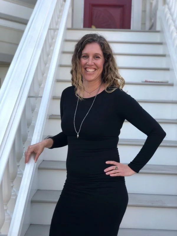 A woman with curly hair wearing a black dress and necklace, smiling on white outdoor stairs in front of a house with red door.