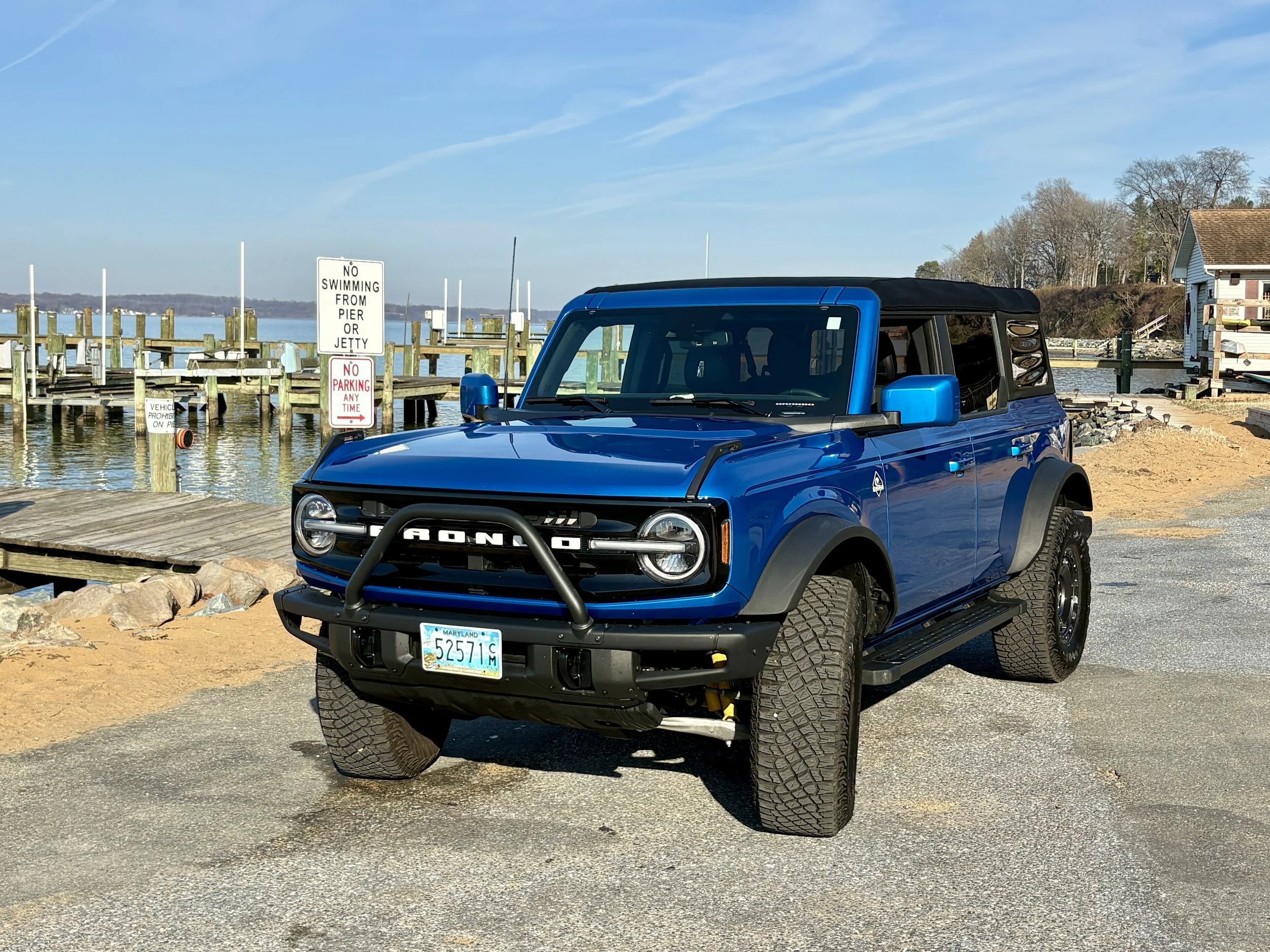 A bright blue off-road vehicle with a Maryland license plate parked near a pier by a calm body of water. The sky is clear with a few wispy clouds, and there are signs indicating no swimming from pier or jetty and no parking at any time.