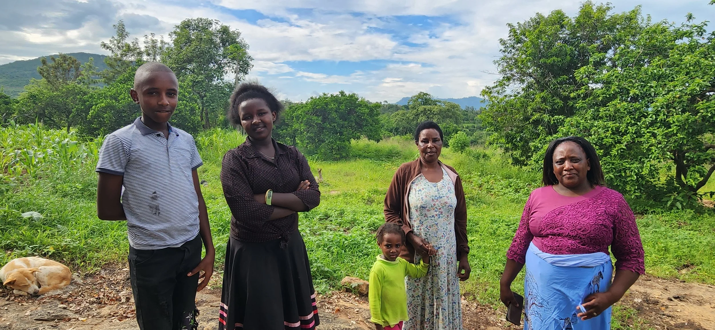 A group of five people, including two children, standing outdoors in a lush, green landscape with trees and mountains in the background. The group includes three women and two children, with a sleeping dog lying on the ground nearby.