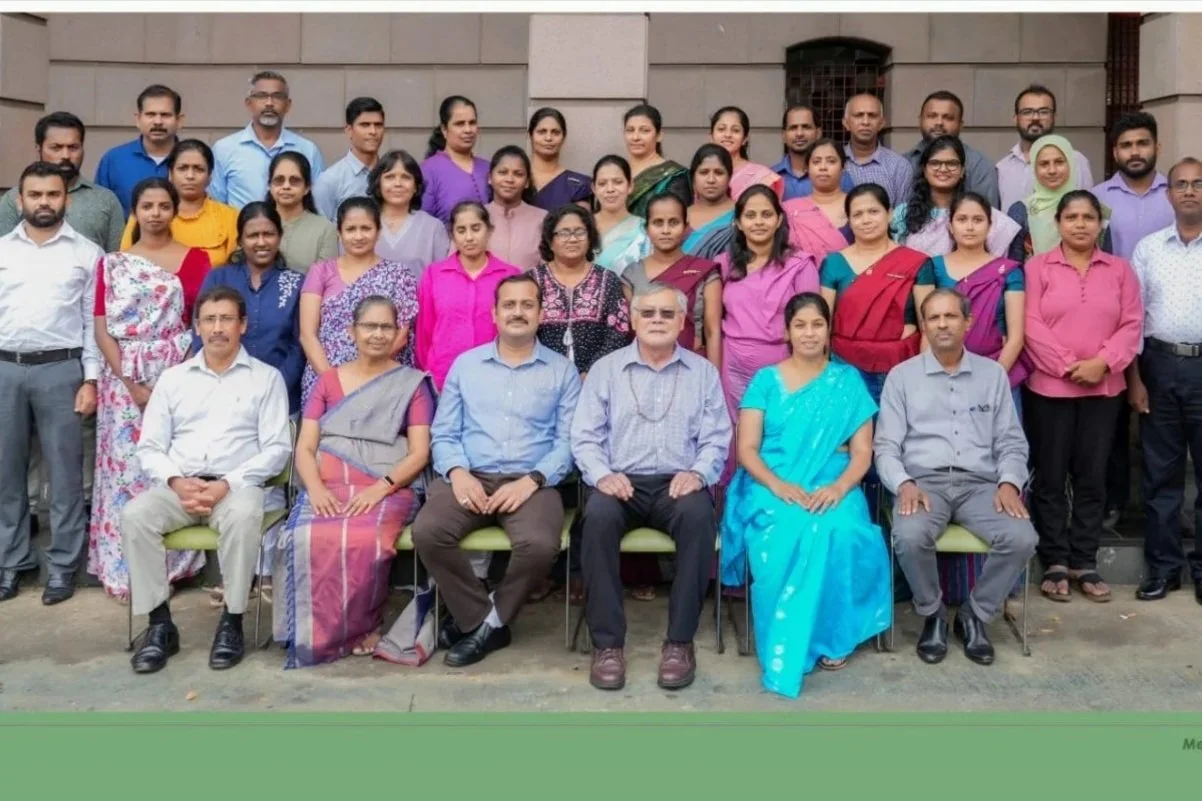 Group of adults posing for a photo outdoors in front of a building, some seated and others standing.