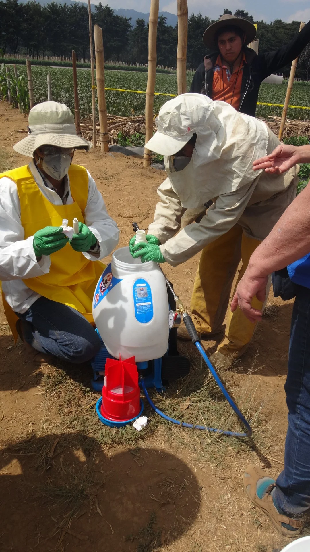 Scientists in protective gear conducting a pesticide application on a farm with crops in the background.
