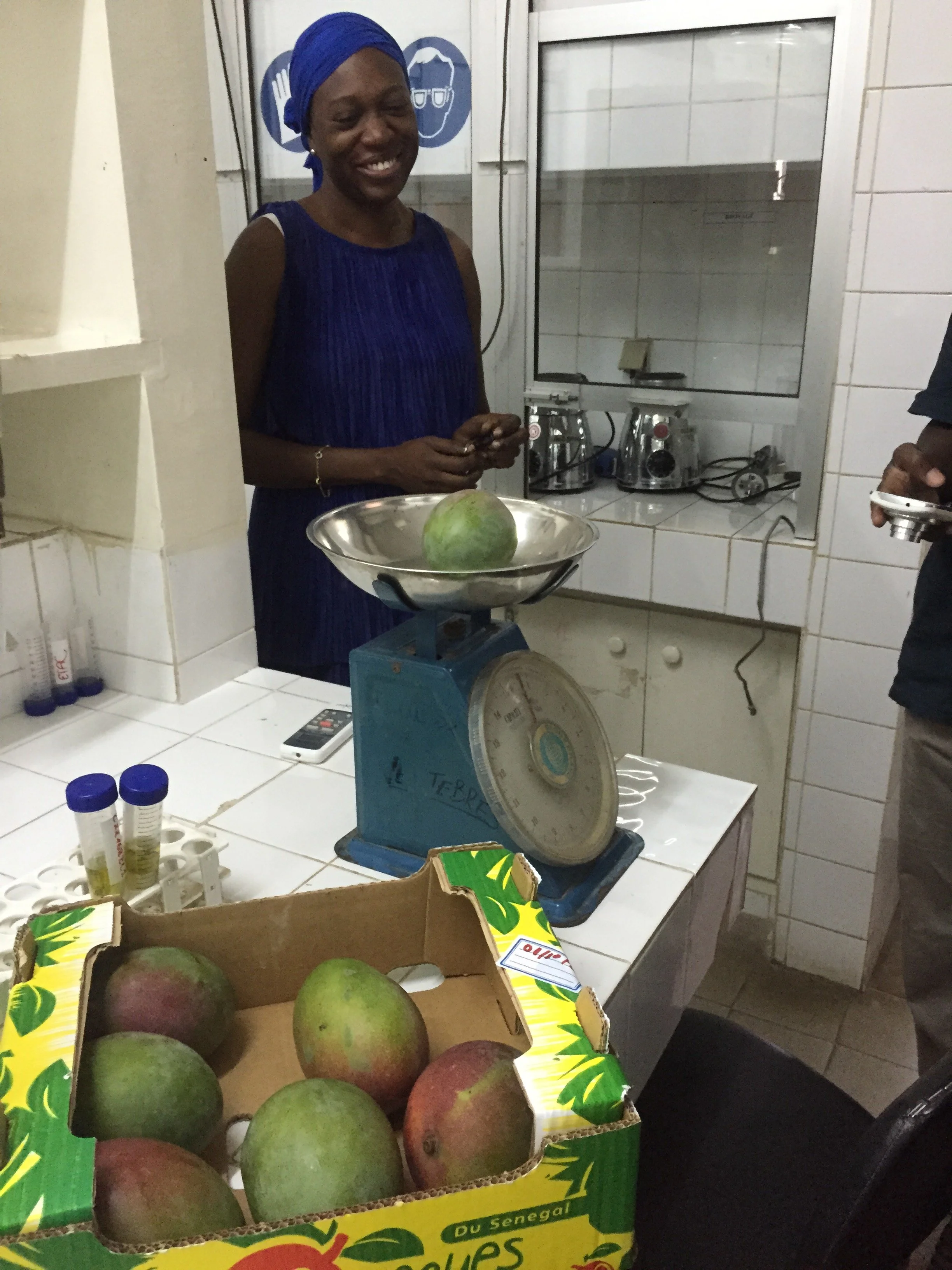 A woman in a blue dress and headscarf smiling in a kitchen with a box of ripe mangoes on the counter, a weight scale, and appliances in the background.
