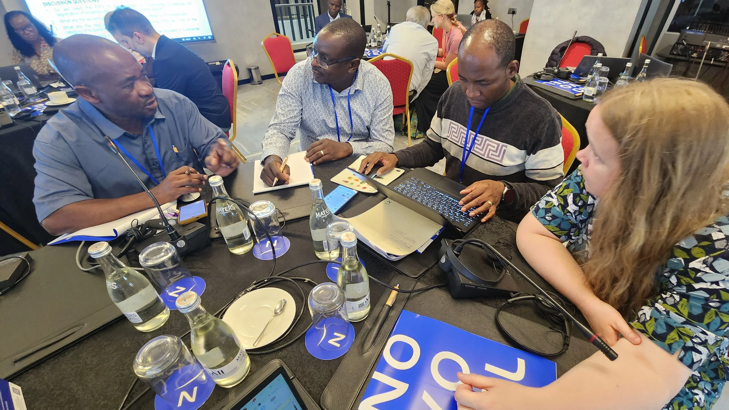 A group of people sitting around a conference table at a professional event, engaged in discussion. There are water bottles, notepads, laptops, and discussion materials on the table. The background shows more attendees and a large screen displaying presentation content.