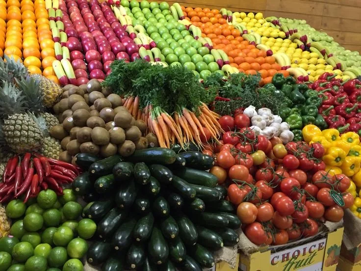 Colorful display of fresh fruits and vegetables, including tomatoes, cucumbers, carrots, garlic, red and green peppers, citrus fruits, and pineapples, arranged in a market stand.
