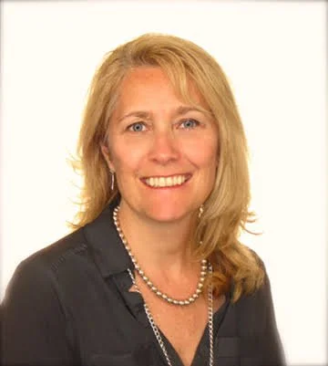 Portrait of a woman with blonde hair, wearing a black shirt, pearl necklace, and earrings, smiling against a white background.