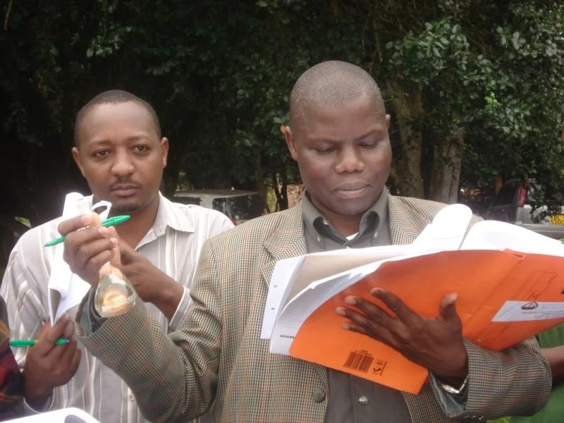 Two men are outside, examining documents. One man is reading a folder, and the other is holding a glass and a pen.