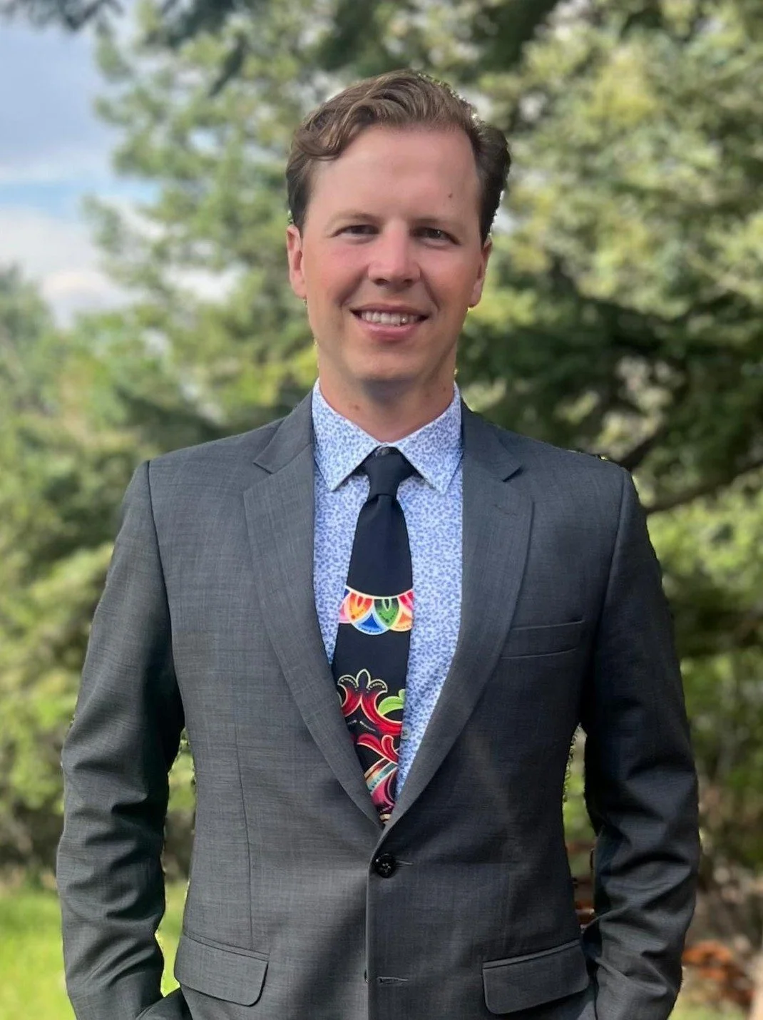 Young man in a gray suit, blue patterned shirt, and colorful tie, standing outdoors in front of trees, smiling.