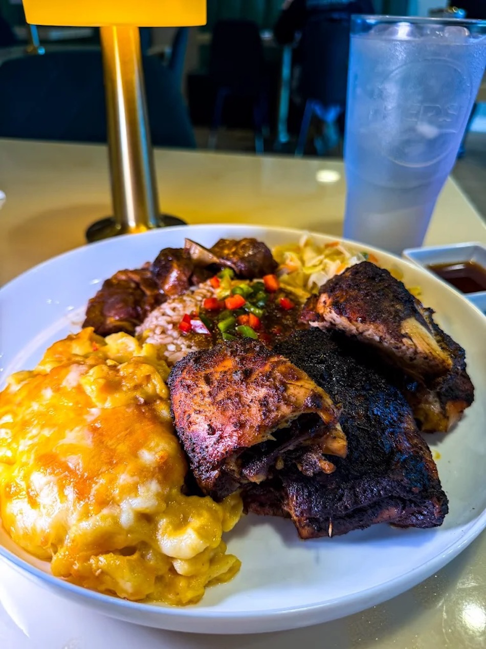 A plate of food featuring barbecued ribs, macaroni and cheese, rice, and grilled vegetables on a table in a restaurant setting.