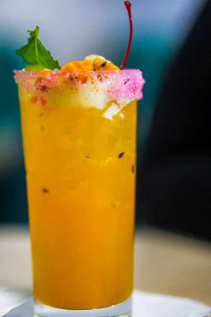 A tall glass of orange-colored beverage topped with pink cotton candy, a cherry, and a green leaf garnish.