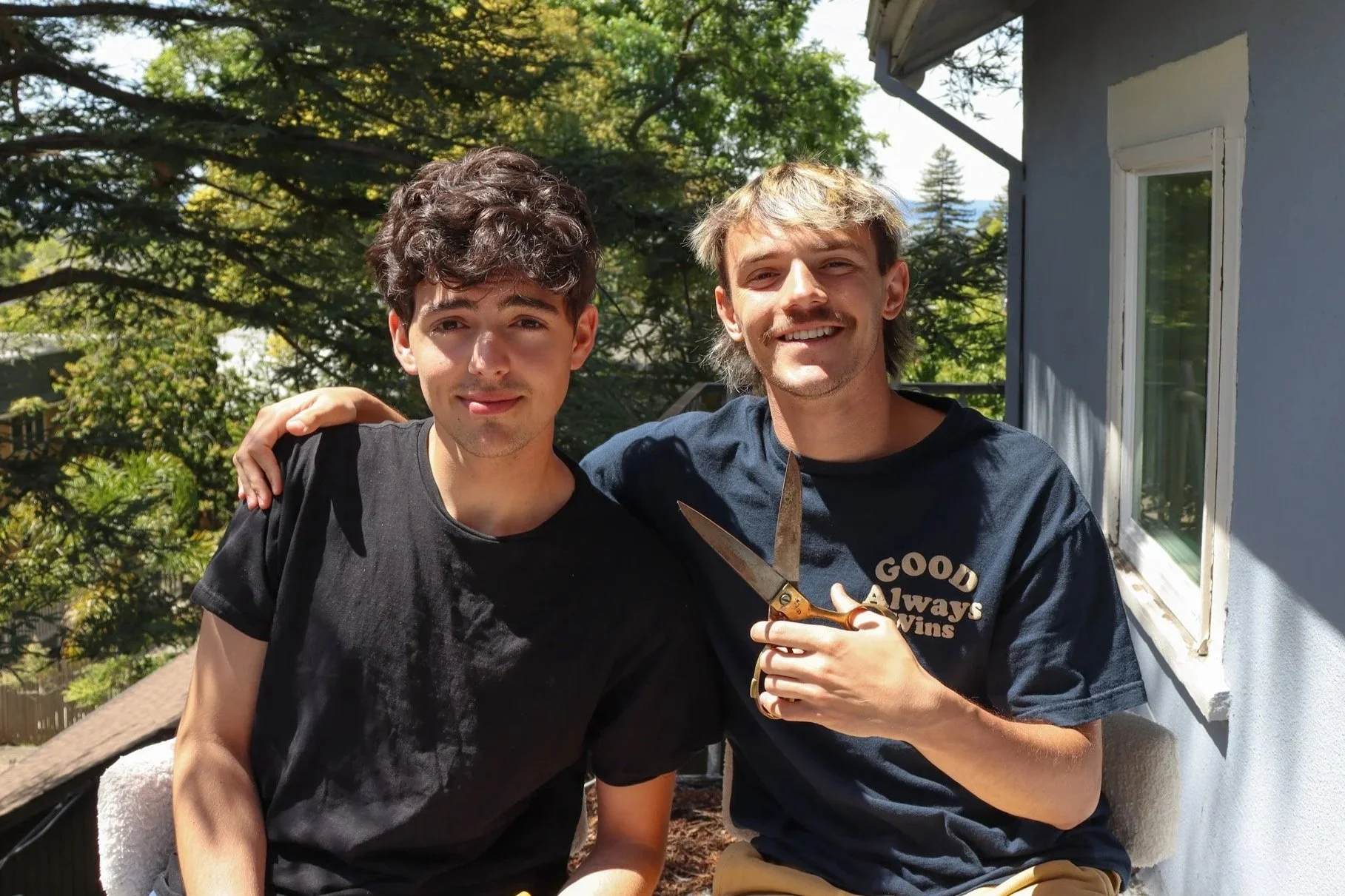 Two young men standing outside on a sunny day, one with curly dark hair wearing a black t-shirt, the other with blonde hair wearing a navy blue shirt with text, holding a pair of scissors, and smiling.