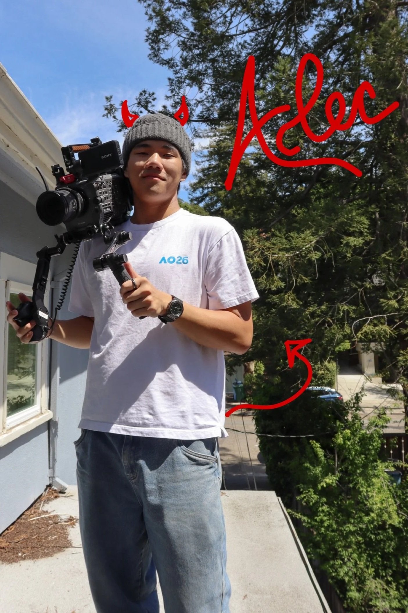 A young man standing outdoors on a sunny day, wearing a gray beanie with red devil horns drawn on it, a white t-shirt, and jeans. He is holding a professional camera on a stabilizer and has a watch on his left wrist. There are trees, a house, and a driveway in the background, with blue sky overhead.