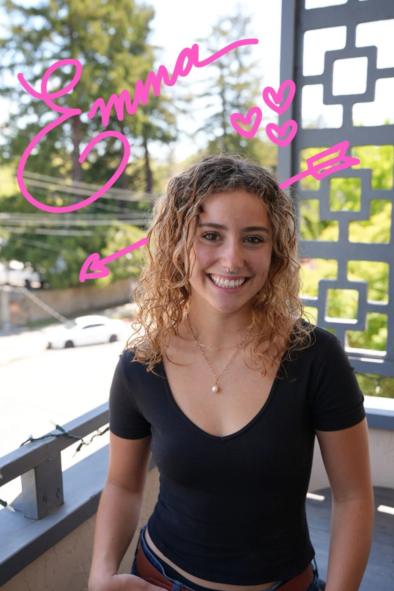 Young woman with curly hair smiling outdoors on a balcony, wearing a black shirt and layered necklaces.