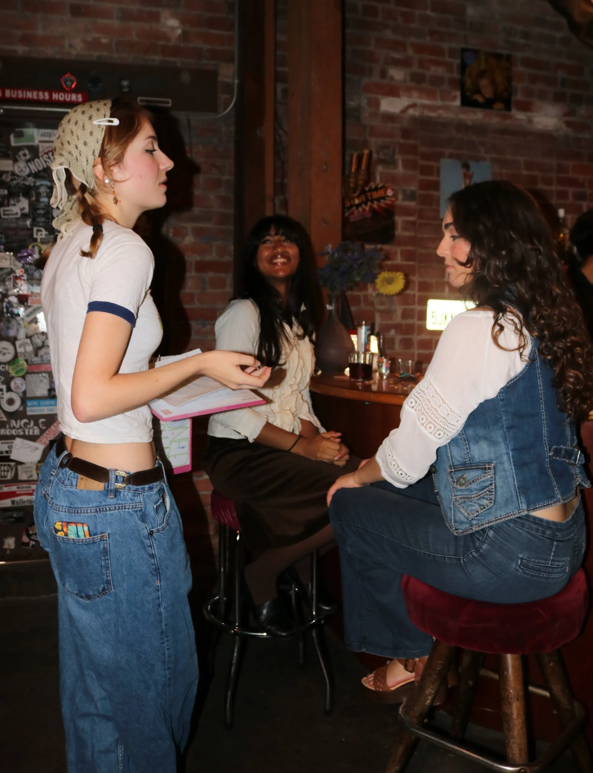 Three women are engaged in a conversation inside a dimly-lit bar with a brick wall background. One woman is standing, another is sitting, and the third is leaning on a high stool. The scene is casual with a cozy atmosphere.