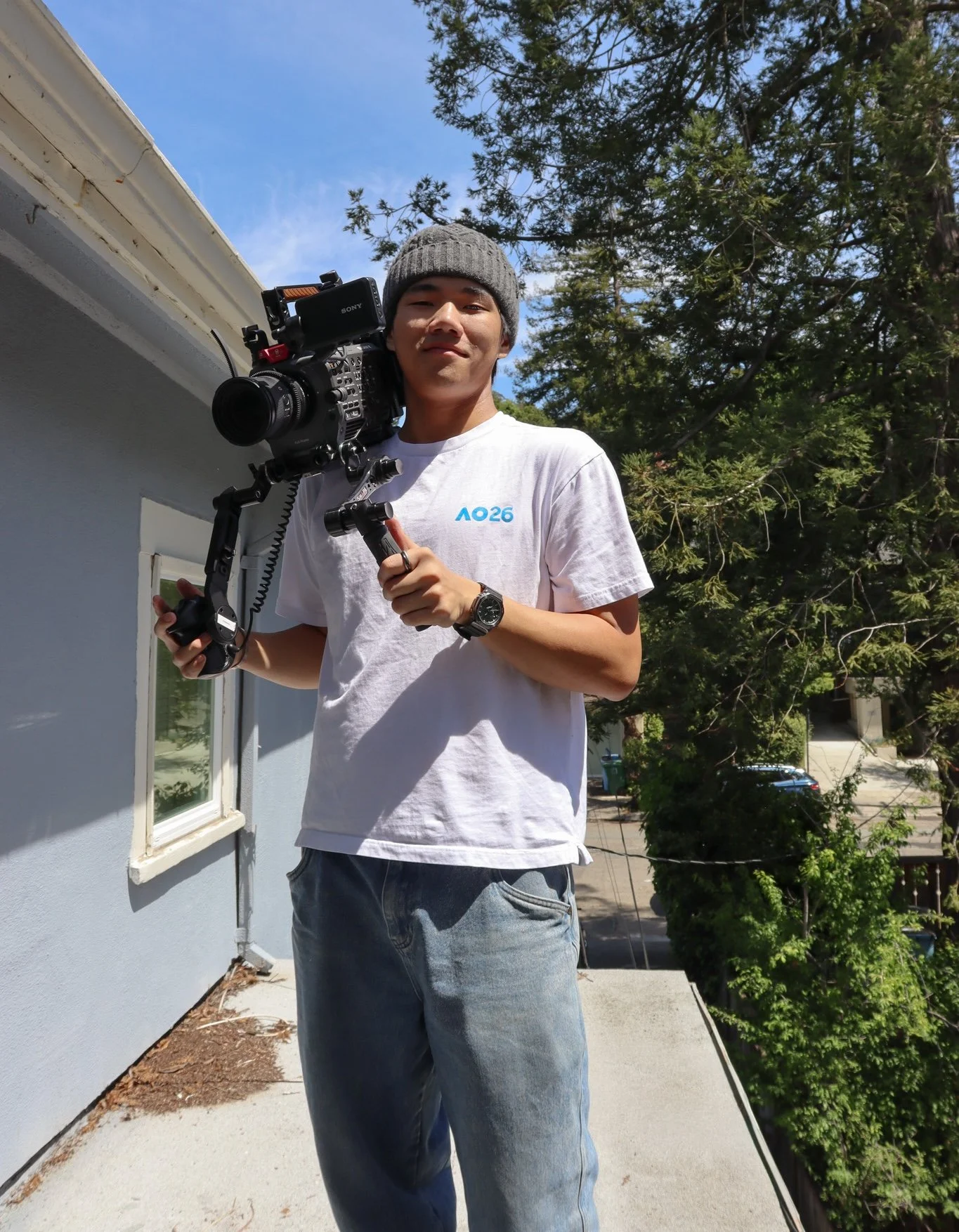 Young man standing outside holding a professional video camera on his shoulder, wearing a gray knit beanie, white T-shirt, and jeans, with trees and a residential house background on a bright sunny day.