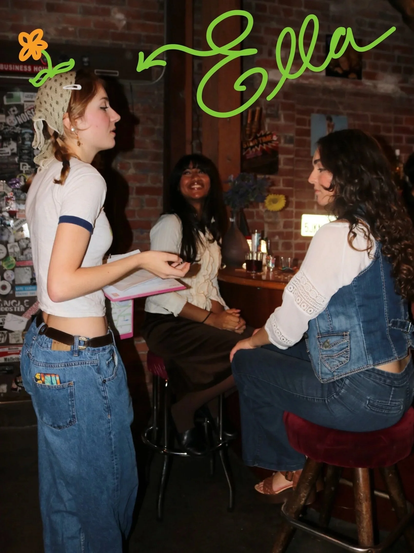 Three women in a bar engaged in conversation. One woman with long, blonde hair in a ponytail wearing a white t-shirt with navy trim and blue jeans holds a notebook. The second woman with curly brown hair, wearing a white blouse and denim vest, is sitting on a barstool. The third woman with long dark hair, dressed in a beige top, is sitting on a red velvet stool, smiling, with drinks and decorations on the bar behind them.