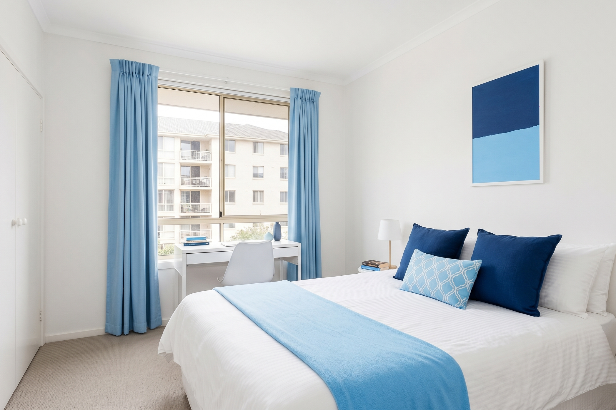 A bright bedroom with white walls, a large window with light blue curtains, a white desk with books, and a bed with blue pillows and a light blue throw blanket.