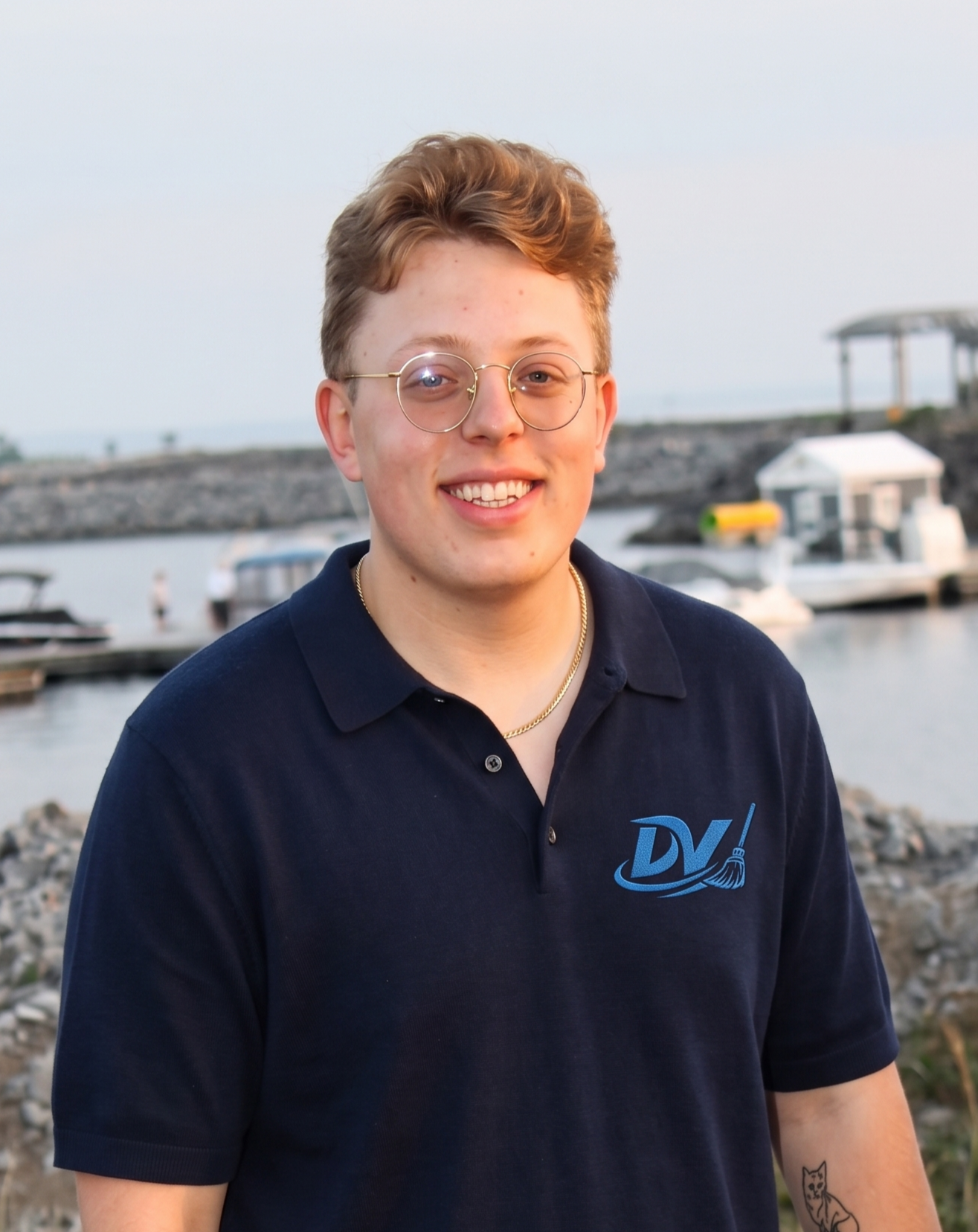 A young man with light skin, short curly brown hair, glasses, and a smile, standing outdoors near a marina with boats and a rocky shoreline in the background.