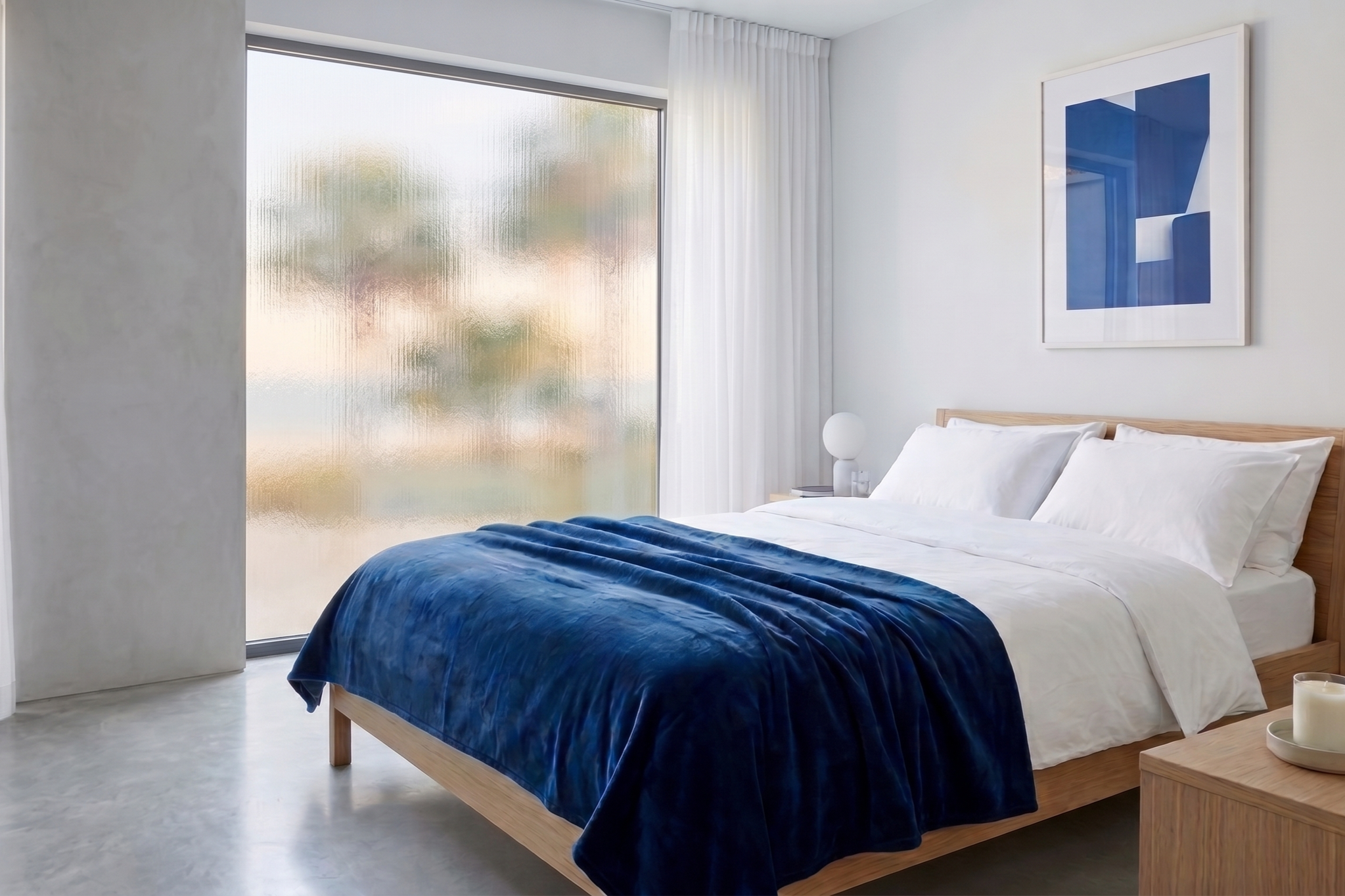 Modern bedroom with a large bed, white bedding, navy blue blanket, on a wooden frame. Tall window with frosted glass and white curtains, minimalist decor including a framed blue abstract art and a small bedside table with a white spherical lamp and a candle.