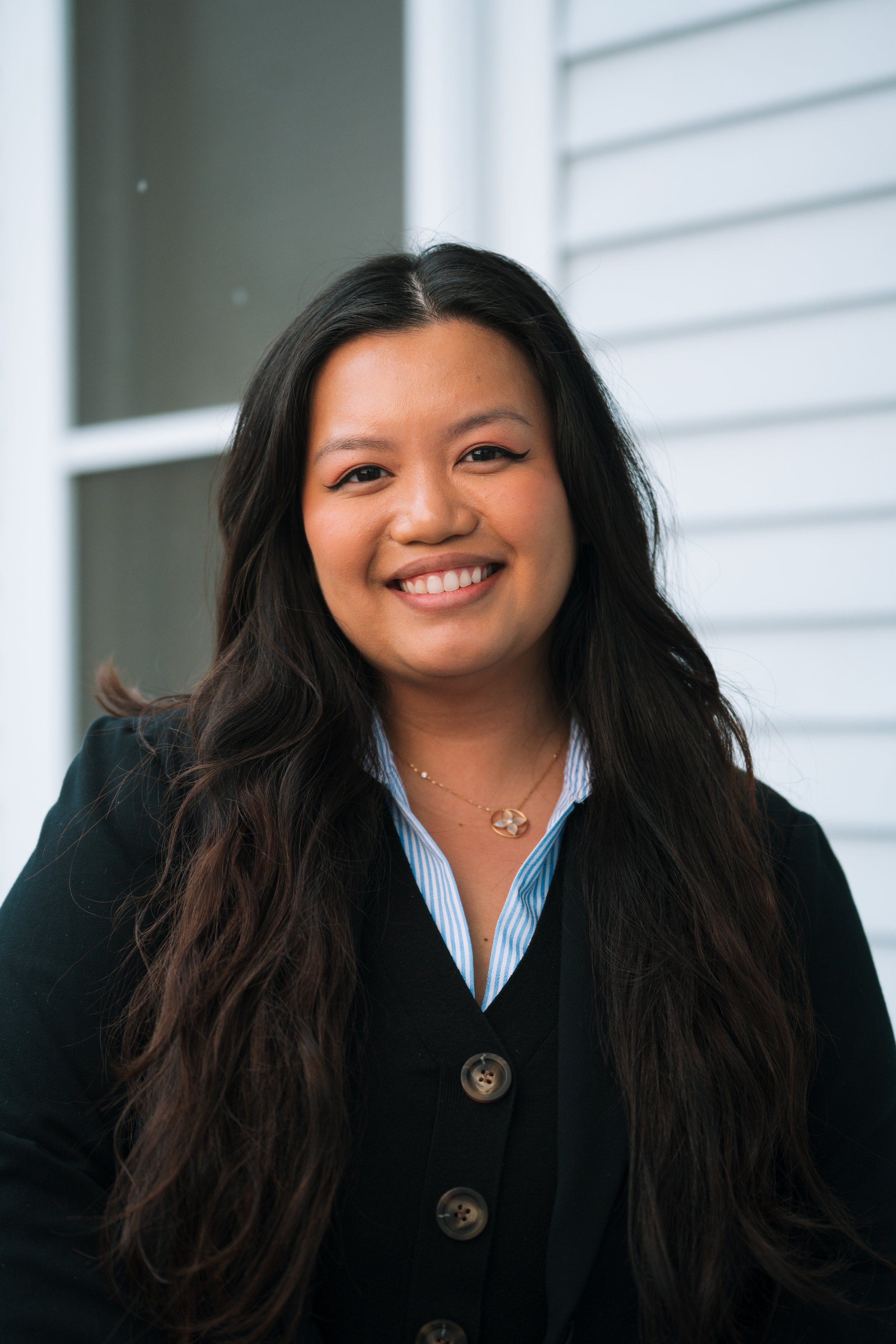 A woman with long, wavy dark hair and a bright smile, dressed in a dark blazer over a blue-striped shirt, standing in front of a window with white blinds.