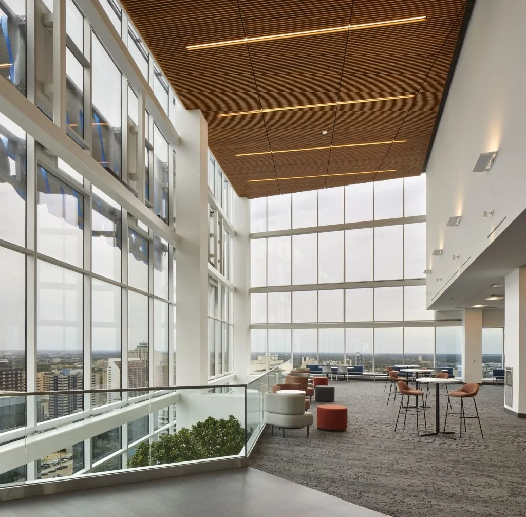 Interior de un edificio moderno con ventanas grandes de vidrio, techo de madera y área de descanso con sillones y mesas.