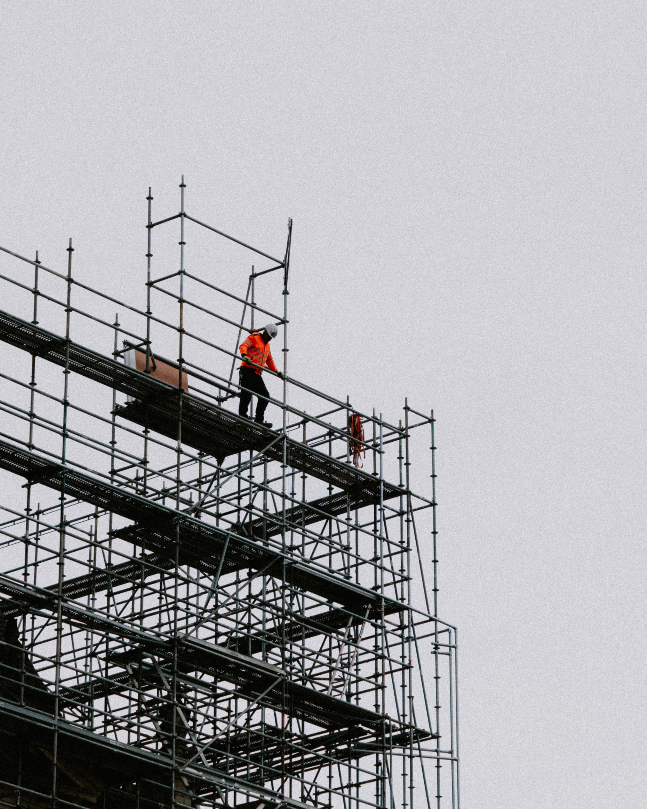 Trabajador en construcción con casco y chaleco naranja en andamios de metal en un cielo gris.
