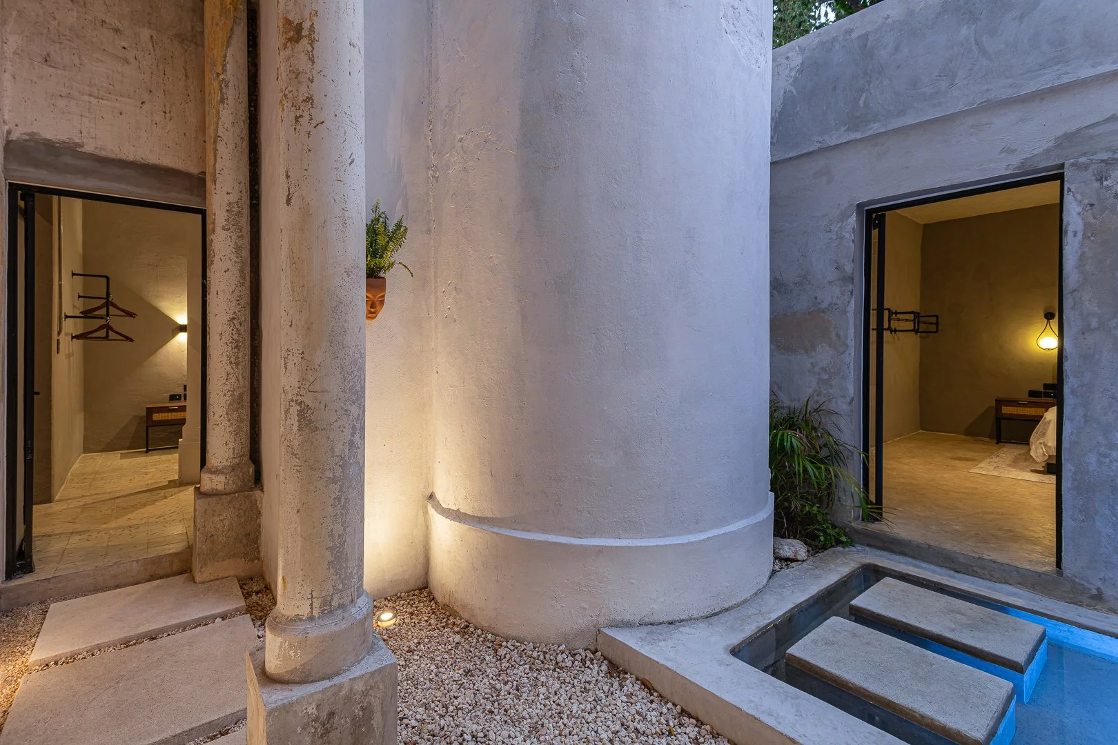 The image shows a modern minimalist courtyard with two rooms visible through open doors, one on each side. The rooms have a warm interior lighting, with one containing a bed and the other a small table and coat hooks. The exterior features a textured