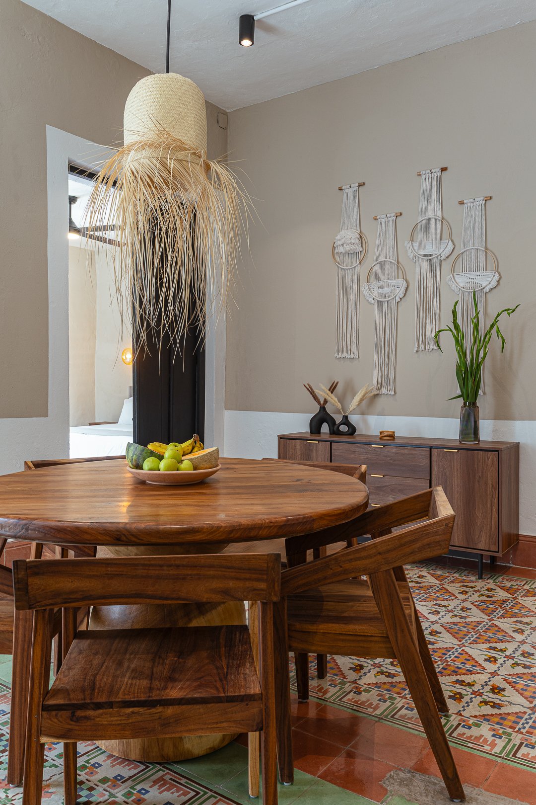 A dining room with a round wooden table, six matching wooden chairs, and a sideboard. The table has a fruit bowl with bananas, apples, and melon. The sideboard is decorated with black vases, dried pampas grass, and a tall green plant in a gray vase. 