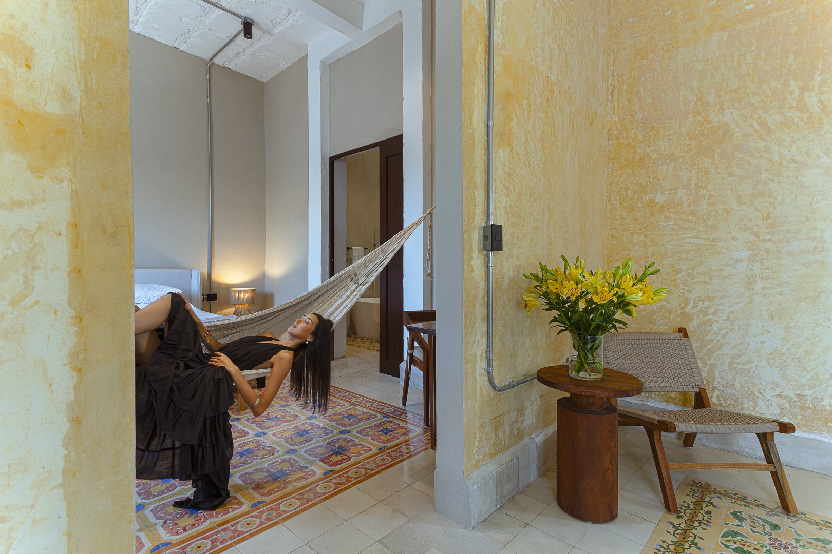 A woman in a long black dress relaxing on a hammock inside a cheerful yellow-walled room with colorful tiled flooring, beside a table with a bouquet of yellow lilies and a transparent chair.