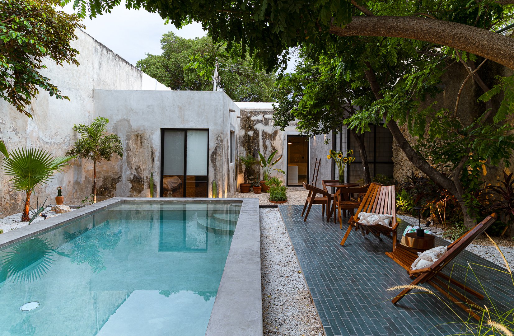 Outdoor patio with a small pool, surrounded by green plants, trees, wooden chairs, and a white weathered wall.