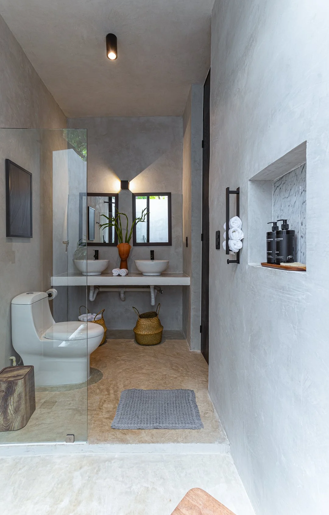 Modern bathroom with two vessel sinks, a toilet, framed mirror above the sinks, a plant in a brown vase, neatly rolled white towels, and bottles of soap or lotion on a wall niche.