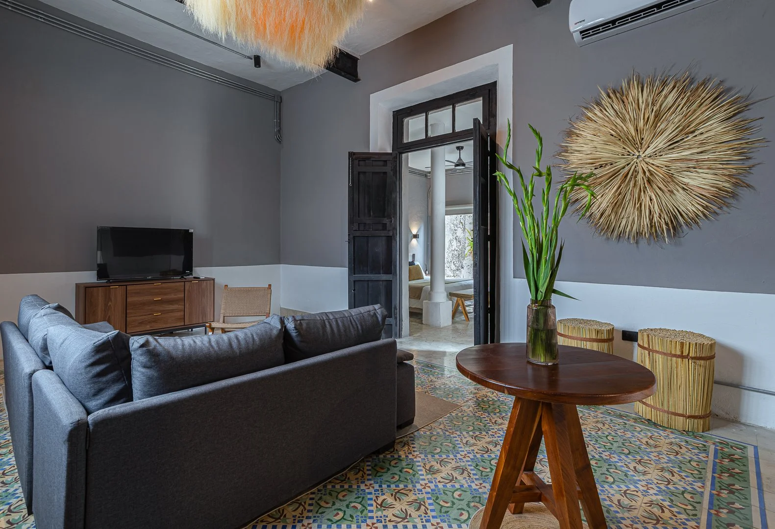 Living room with gray sofas, a wooden TV stand with a TV, a round wooden table with a vase of green bamboo, patterned tile flooring, decorative wall art, and a doorway leading to a bedroom.