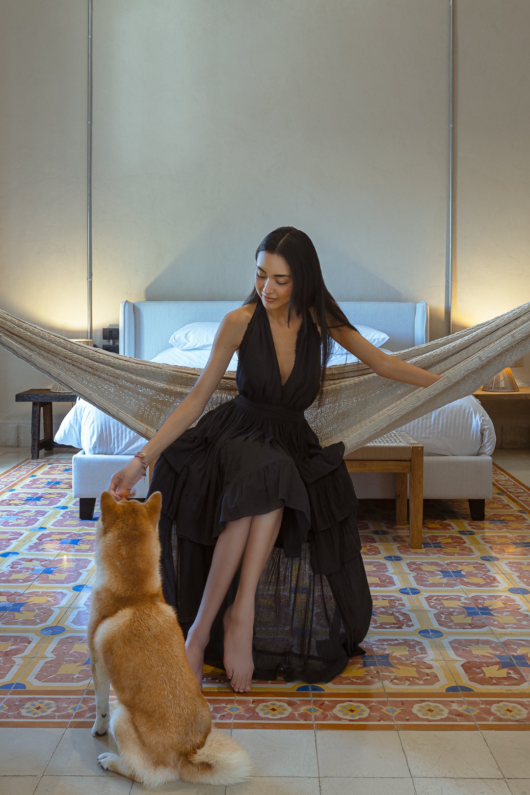 A woman in a black dress sitting on a bed, petting a sitting dog in a well-lit hotel room with colorful tiled floor.