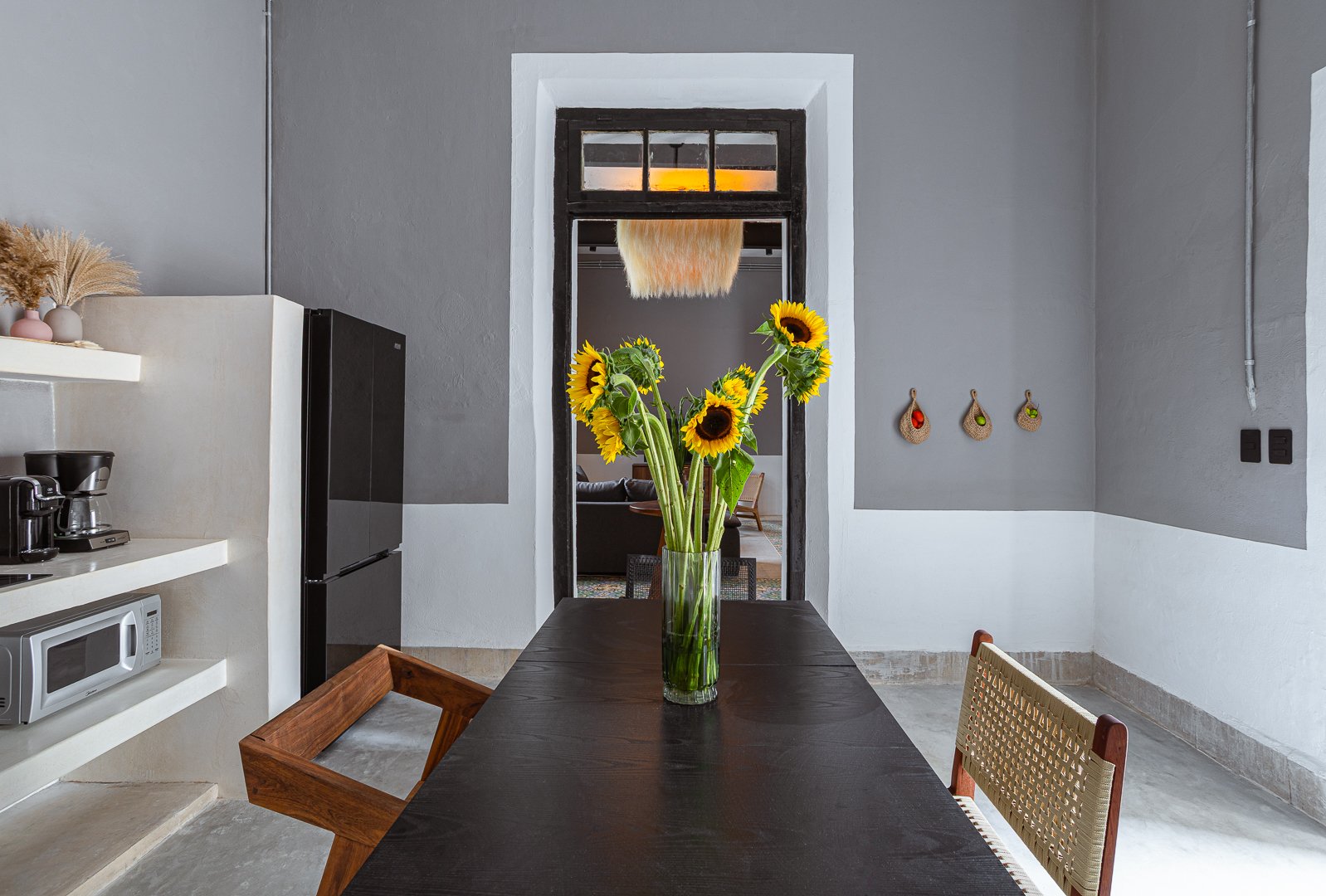 A dining room with a dark wooden table, a glass vase of yellow sunflowers, and a woven chair. The background has a doorway showing a living room with a black sofa and a hanging light fixture. The wall to the right has three small, decorative hanging 