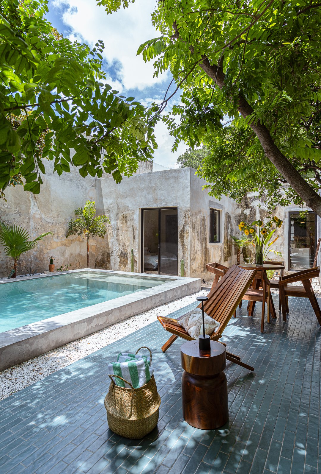 A backyard patio with a small pool, wooden lounge chair, round wooden side table, and a table with a vase of yellow flowers. Surrounded by greenery, including trees and potted plants, with a rustic wall and partly cloudy sky overhead.