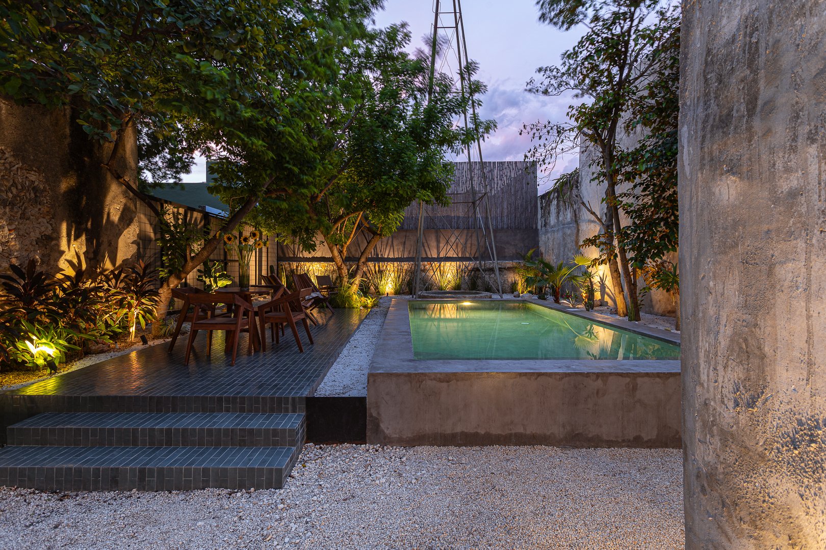 A backyard with a small pool, outdoor seating, trees, and decorative lighting at dusk.