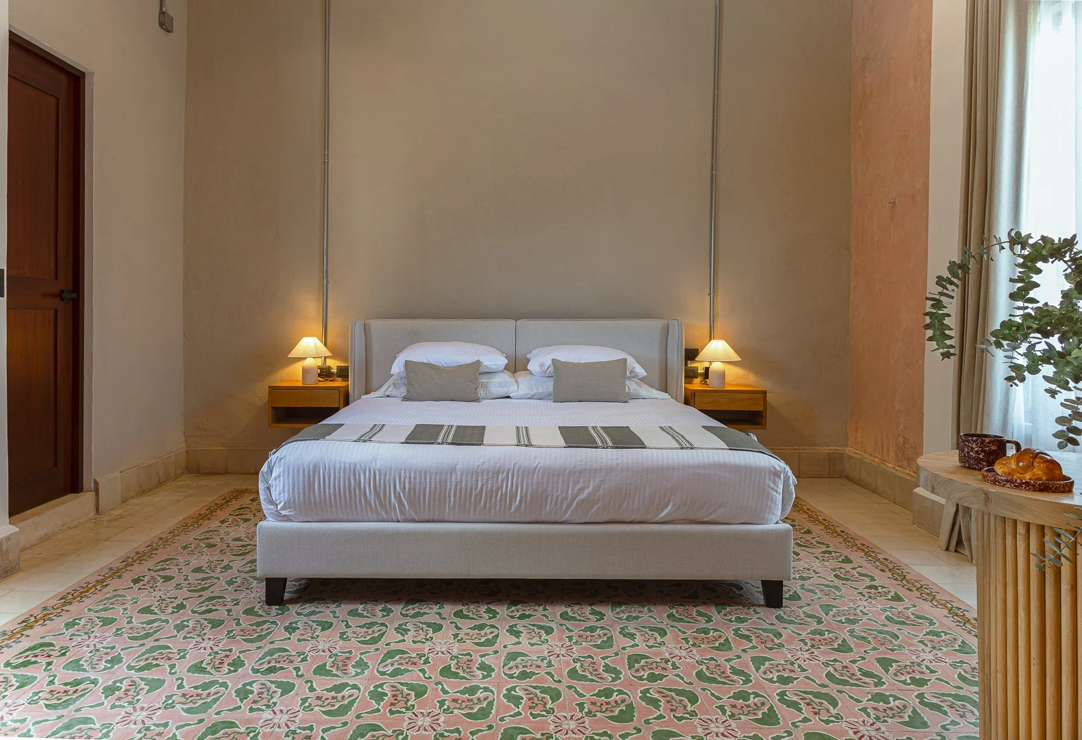A neatly made bed with white linens, two pillows, and two smaller beige pillows in a hotel room. There are two wooden nightstands with lamps on each side of the bed. The room has beige and peach-colored walls, a patterned rug, and a small table with 