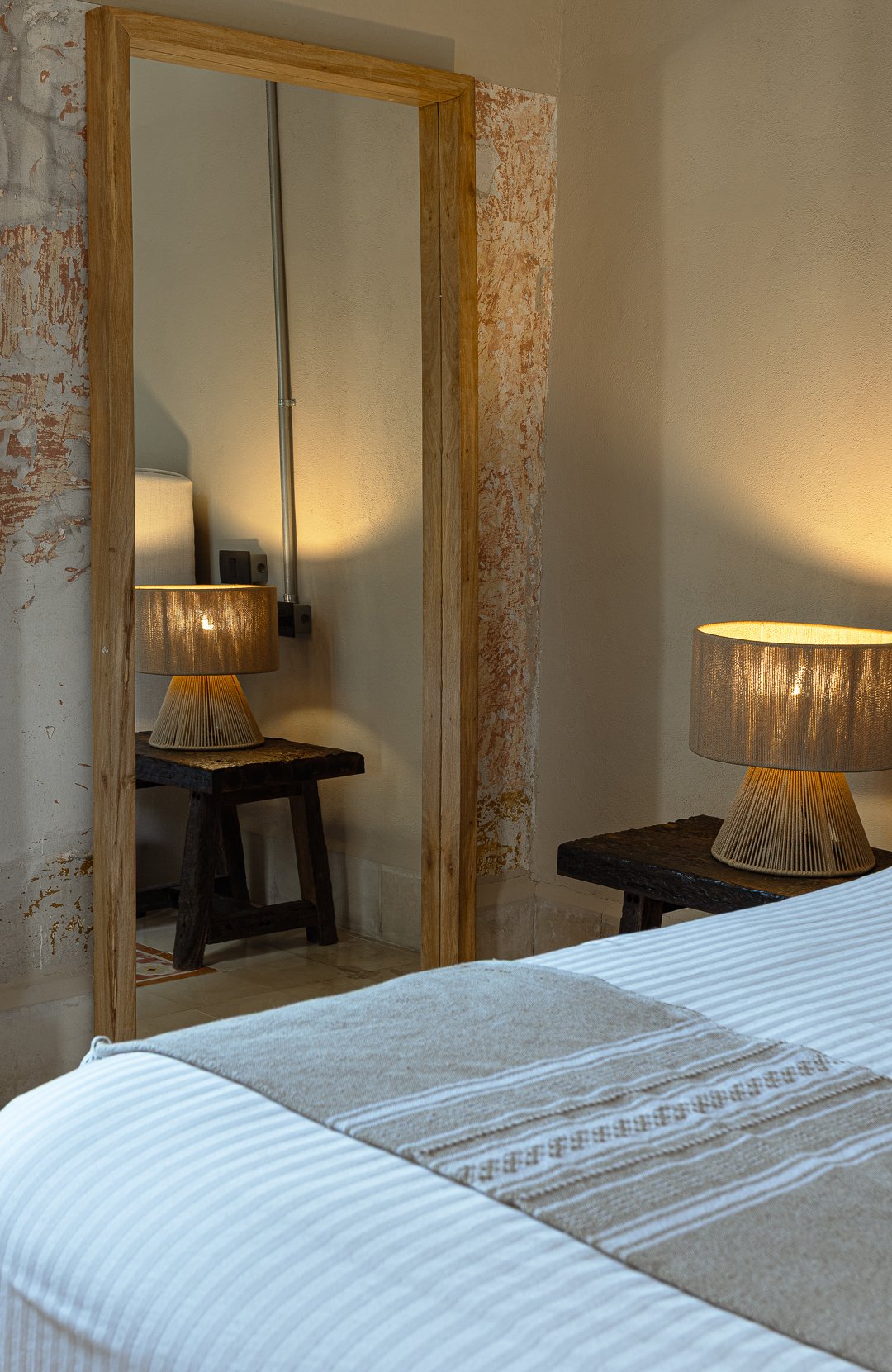 A bedroom with a bed, a wooden nightstand, and a table lamp with a beige lampshade. A mirror with a wooden frame reflects part of the room, showing another nightstand and lamp. The room has a distressed wall with exposed plaster patches.