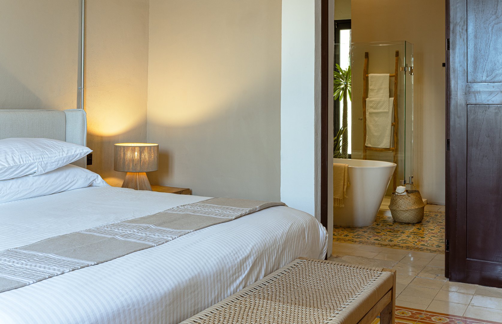 A bedroom with a bed, pillows, a nightstand, and a lamp. A bathroom with a bathtub, a towel rack with towels, and decorative floor tiles is visible through an open door.